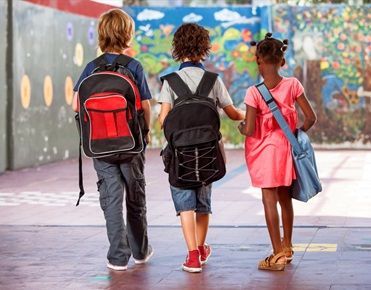 Trois enfants portant des cartables ou sac à dos sont vus de dos dans la cour de leur école