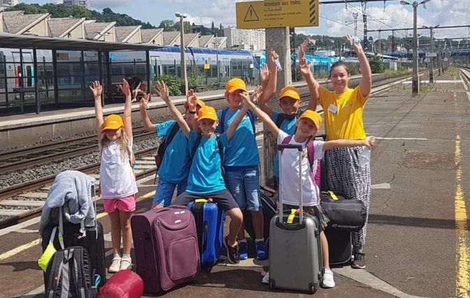 Des enfants lèvent la main sur un quai de gare, accompagnés par une animatrice