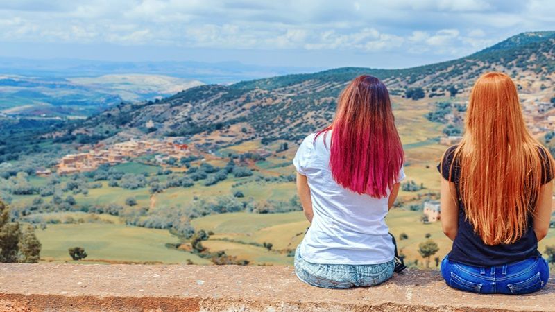 Deux jeunes filles de dos sont assises sur un muret et contemplent la vue de colines