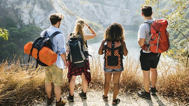 Des jeunes gens, sac à dos et toile de tente sur le dos, admirent une crique depuis un escarpement rocheux