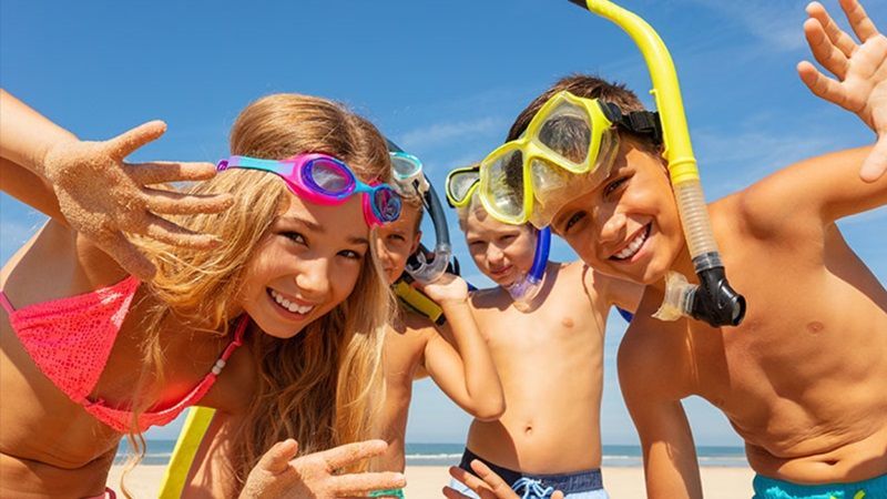 Des enfants, portant un masque et un tuba, sourient et prennent la pose devant un ciel bleu