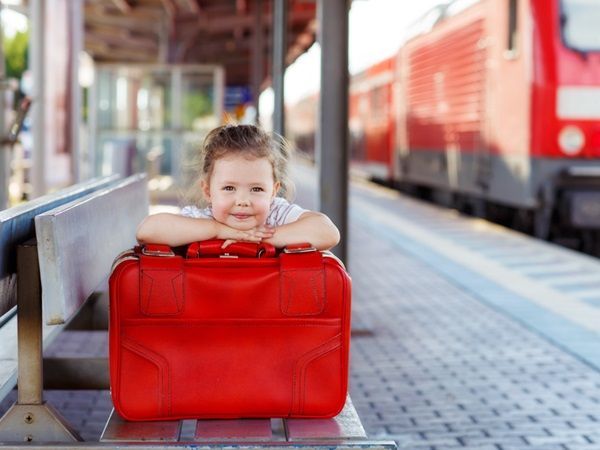 Une petite fille est accoudée sur sa valise rouge sur le quai d'une gare avec un train en fond