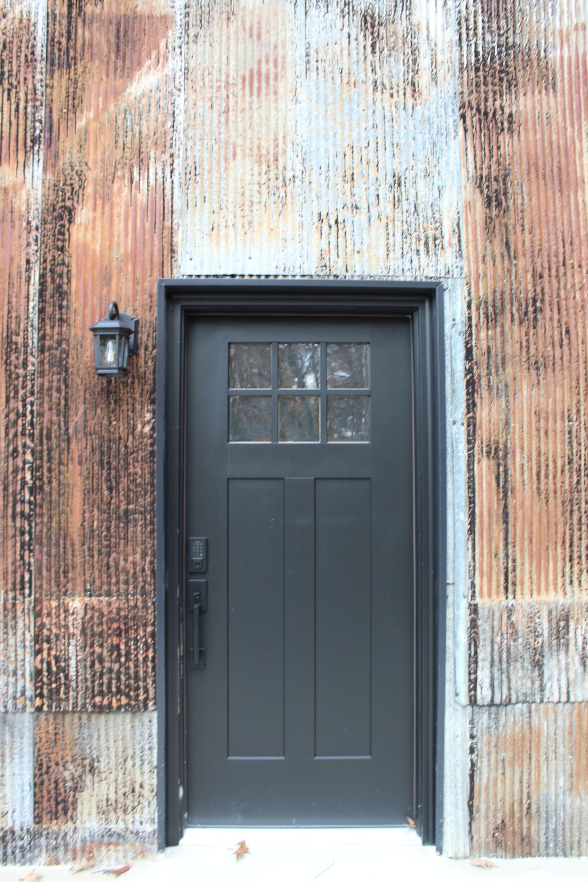 A black door is sitting on a rusty metal wall with a key pad entry opening to a garage area connected to the kitchen.