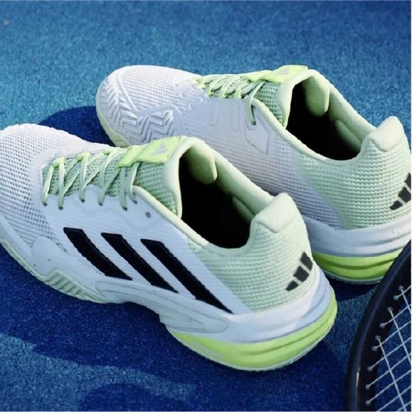 Scarpe da tennis Adidas bianche con strisce nere e dettagli verdi su una superficie blu.