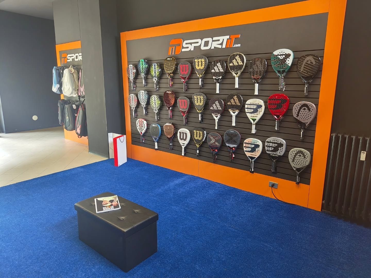 Esposizione di racchette da paddle su una parete all'interno di un negozio di articoli sportivi con moquette blu e pareti nere.