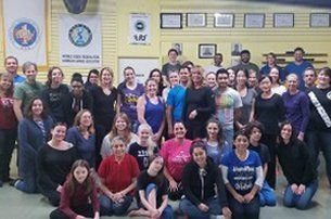 People of varying ages in casual clothing stand and kneel in front of a yellow wall.