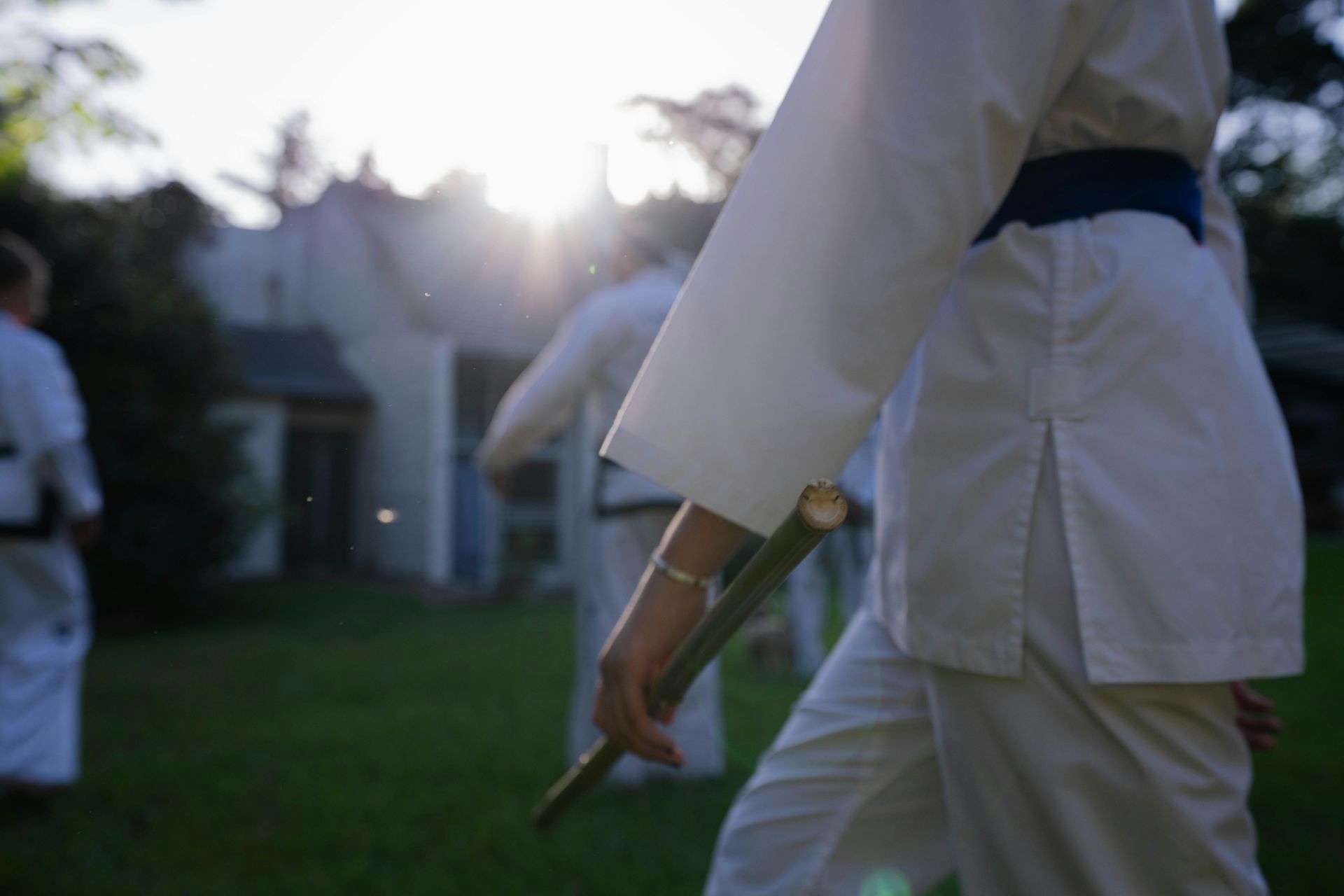 Person in white martial arts uniform holding sticks, outside, in front of others.