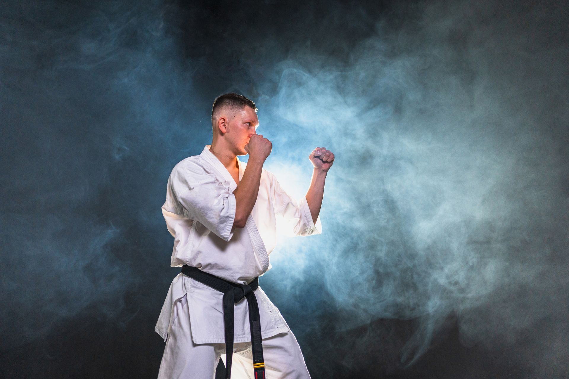 Karateka in a white uniform, black belt, in fighting stance, with fists up, against smoky background.