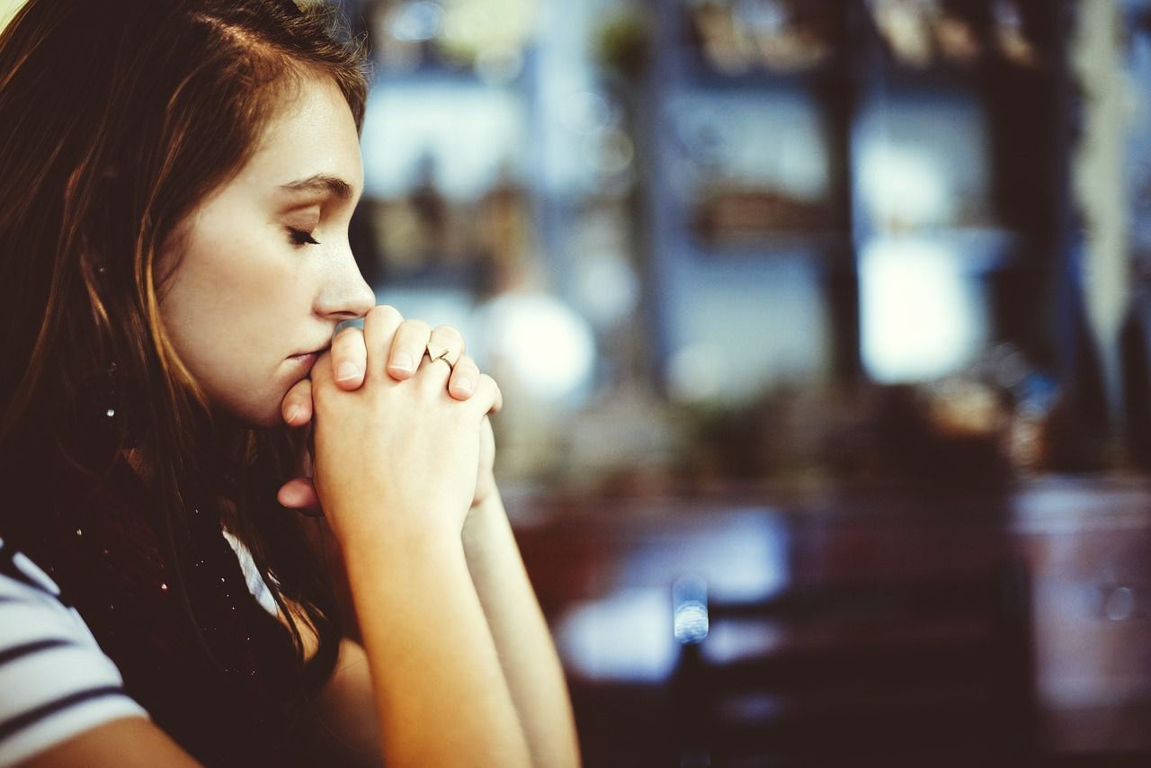 A young woman is sitting at a table with her hands folded in prayer.