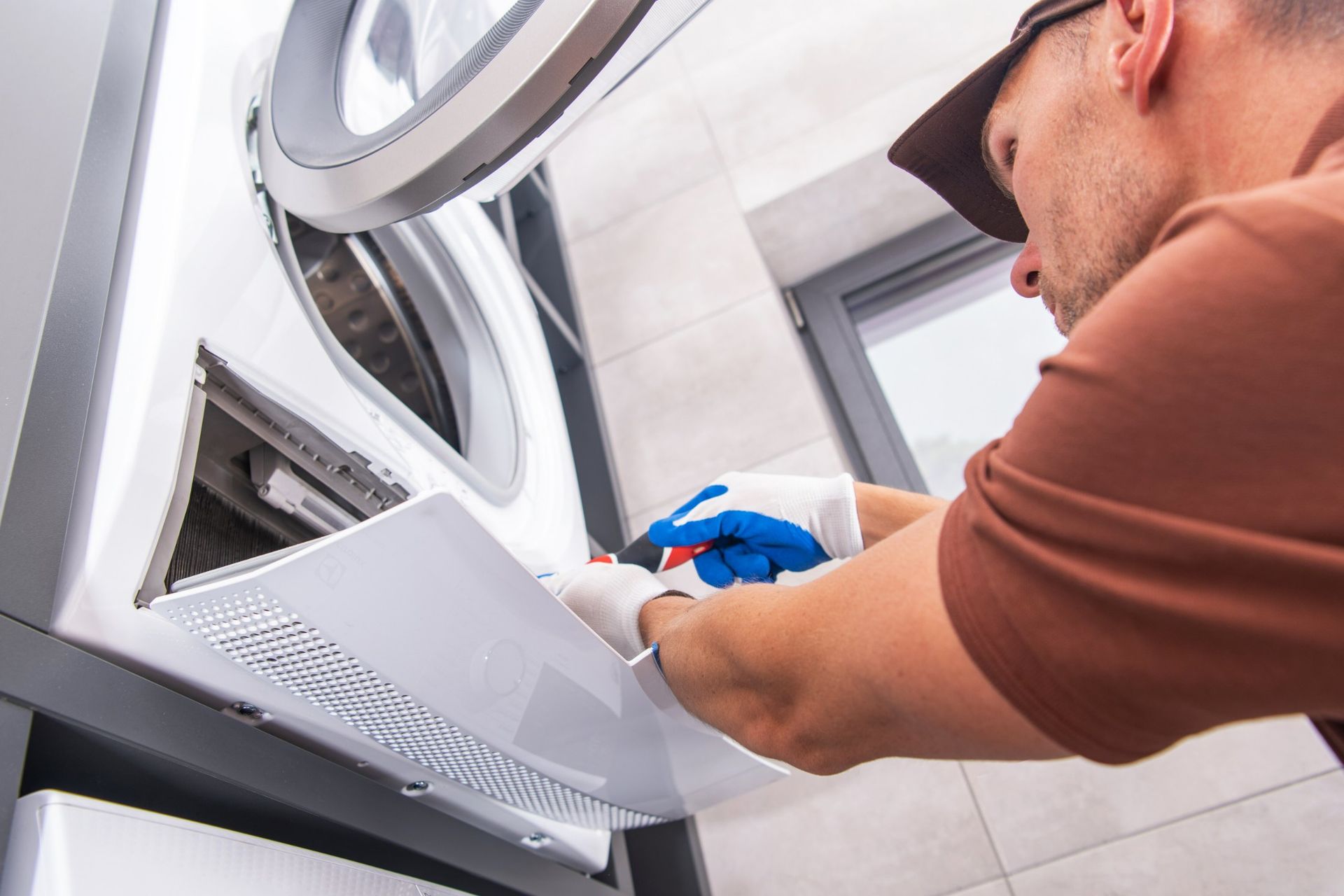 Man in gloves fixing a white washing machine; indoors, near a window.