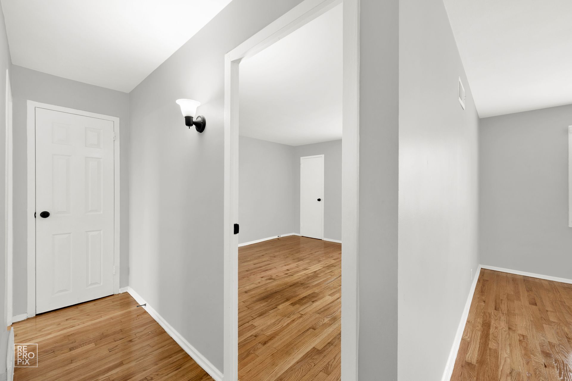 A hallway with white doors and light grey walls. A room with hardwood floors is visible through an open doorway.