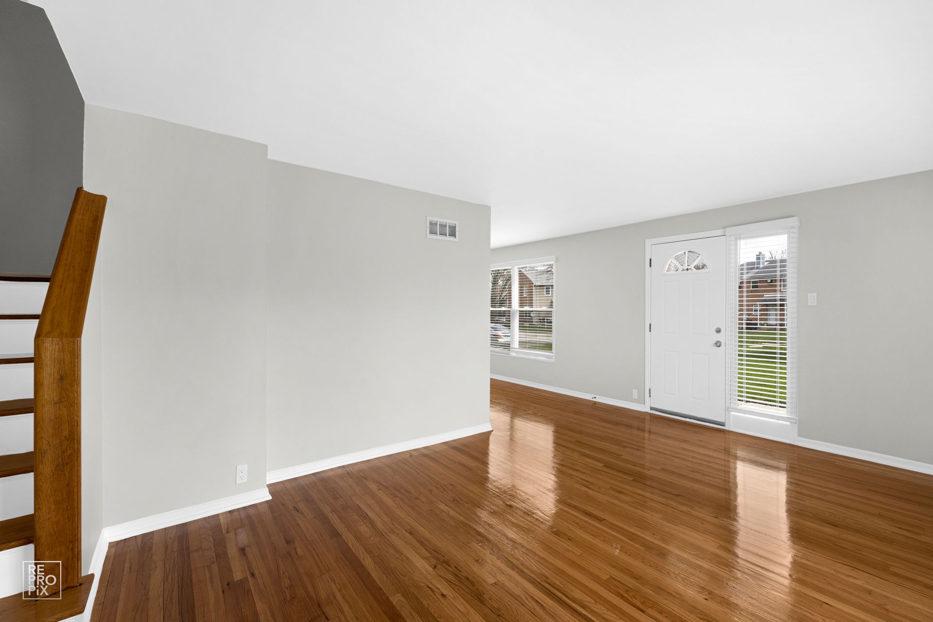 Empty living room with hardwood floors, gray walls, stairs, and front door with sidelight.