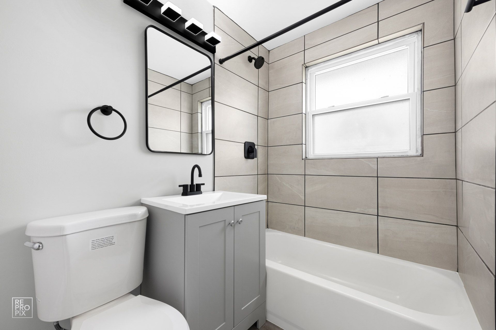 Bathroom with gray vanity, toilet, and tiled walls. Black fixtures and a window.