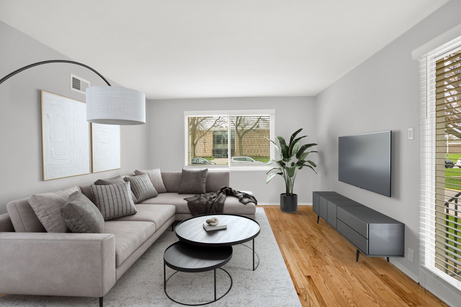 Modern living room with gray sectional sofa, TV, and coffee tables. Light wood floors, neutral walls, and a large window.