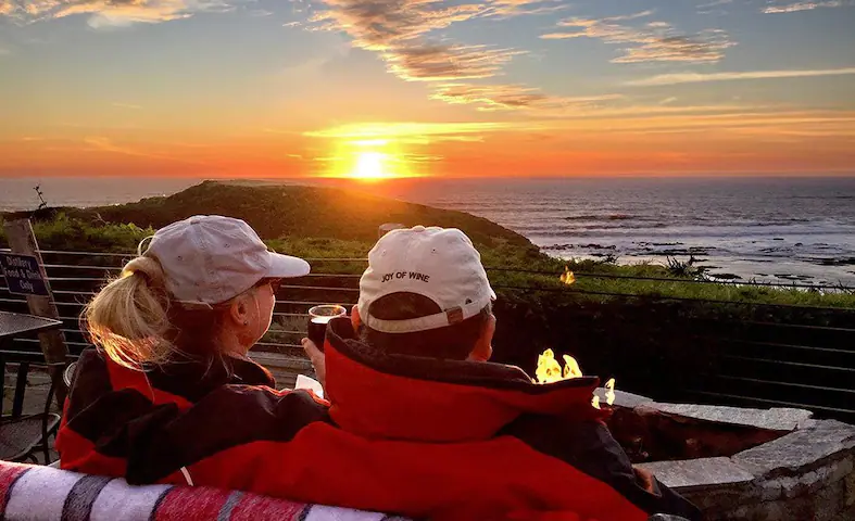 Una pareja sentada en la terraza de Moss Beach Distillery disfrutando de una copa de vino y la vista del atardecer al mar.