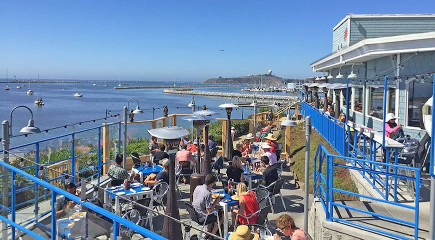 Crowded sunny Sam's Chwder House terrace overlooking Pilar Point Harbor. People are eating.