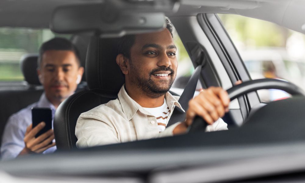 A man is driving a car with another man sitting in the back seat.