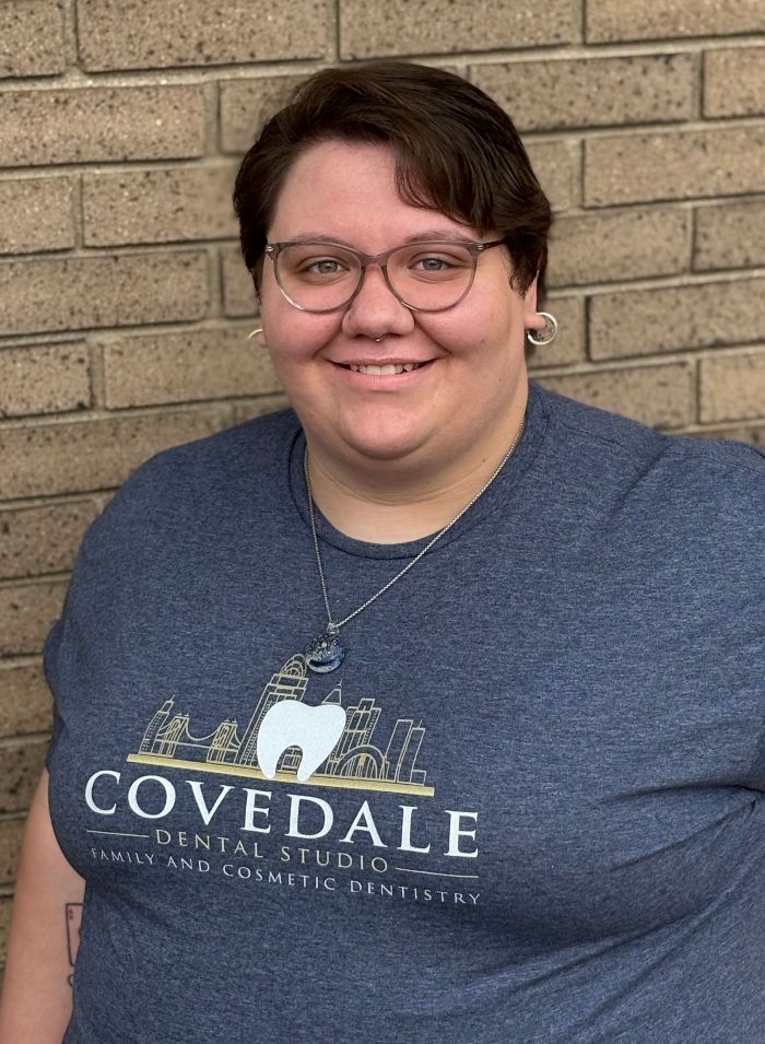 A woman wearing glasses and a blue t-shirt is smiling in front of a brick wall.