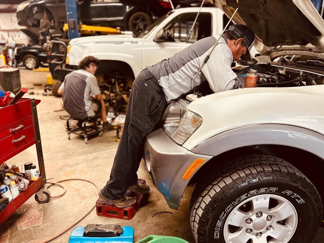 Two mechanics working on vehicles in a garage. One leans over a car engine; the other works on a vehicle underneath.
