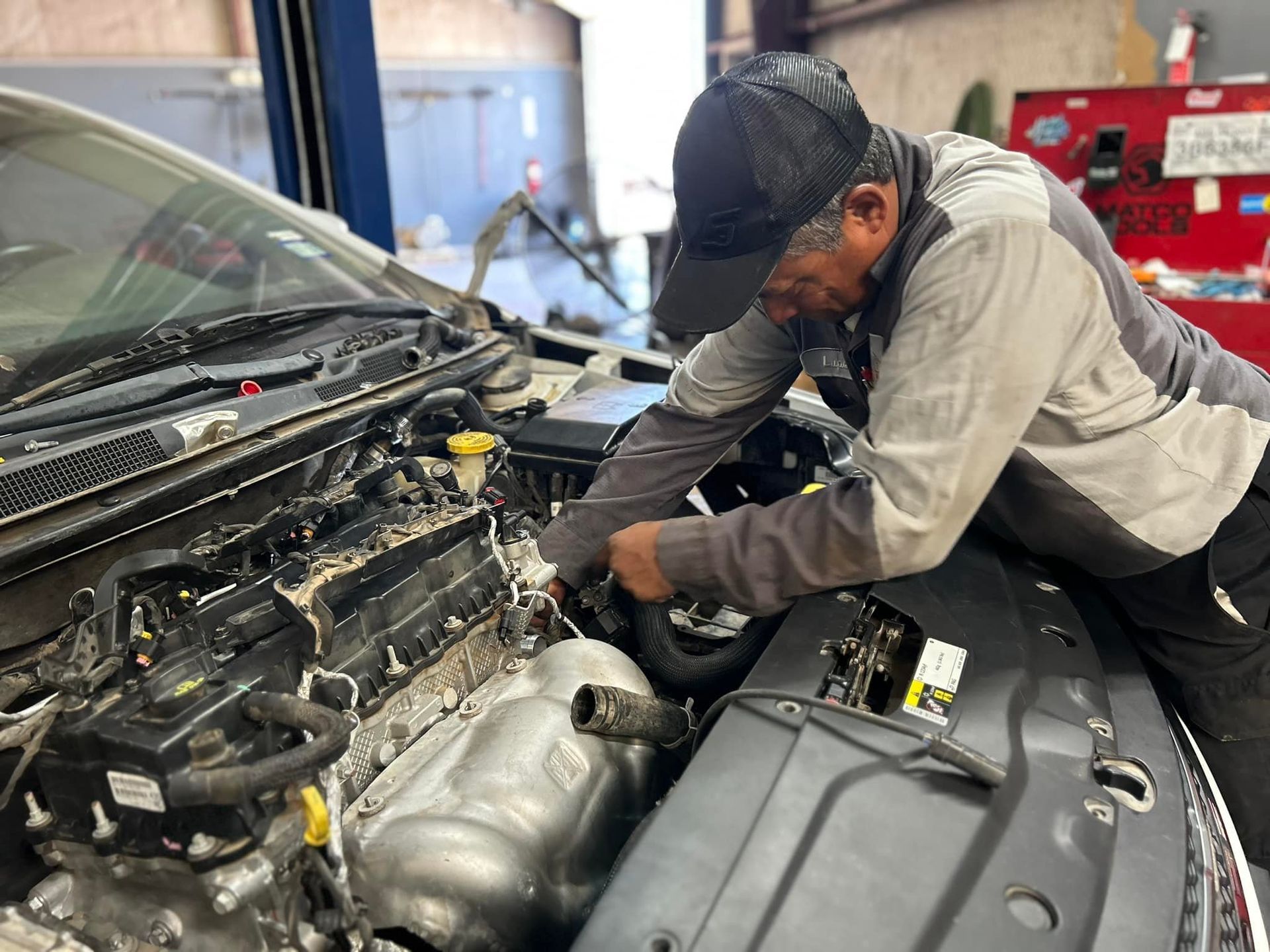 Mechanic working on a car engine, wearing a cap and gray uniform, inside a garage.