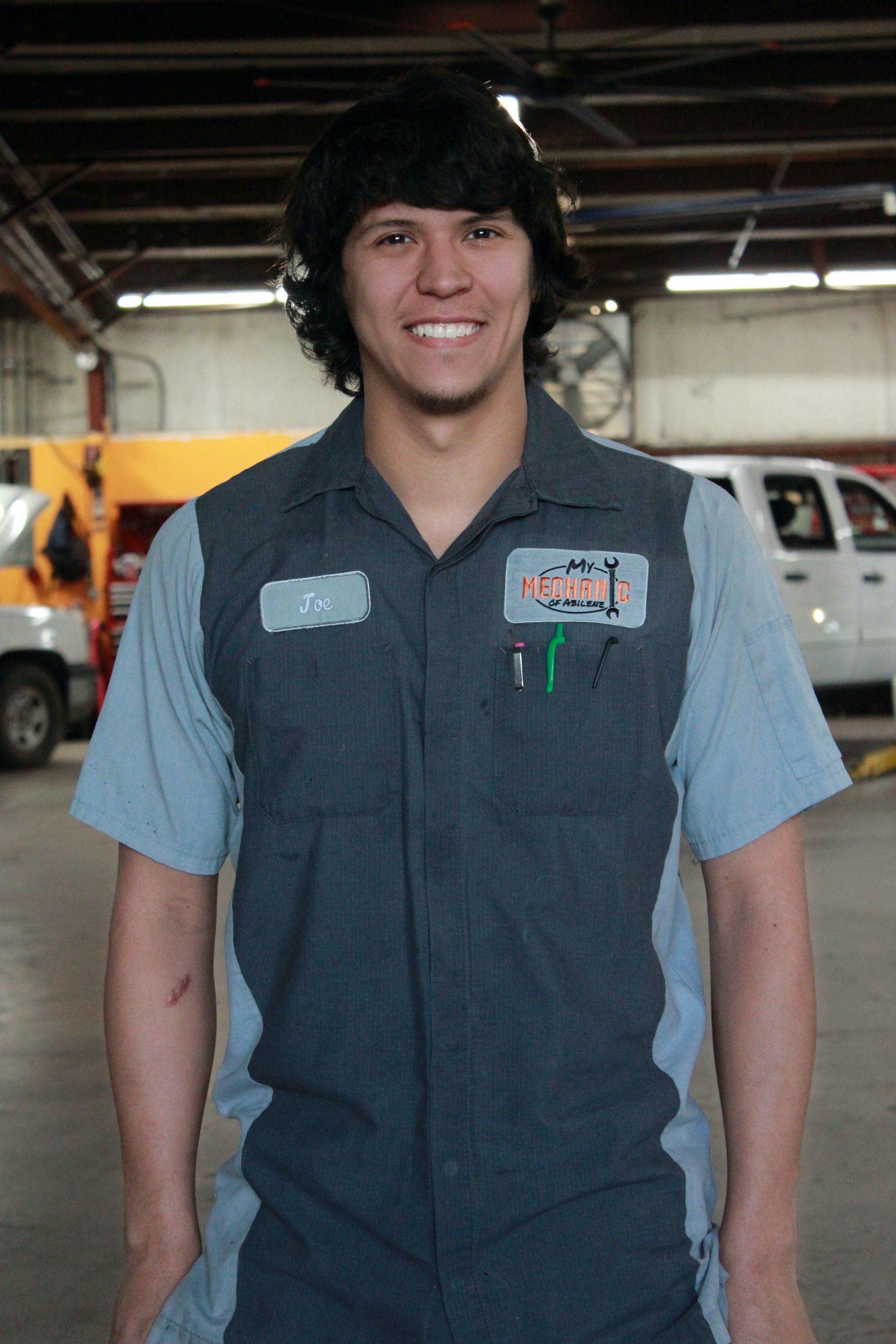 Mechanic in grey and blue uniform smiles in a garage; a truck and equipment are in the background.