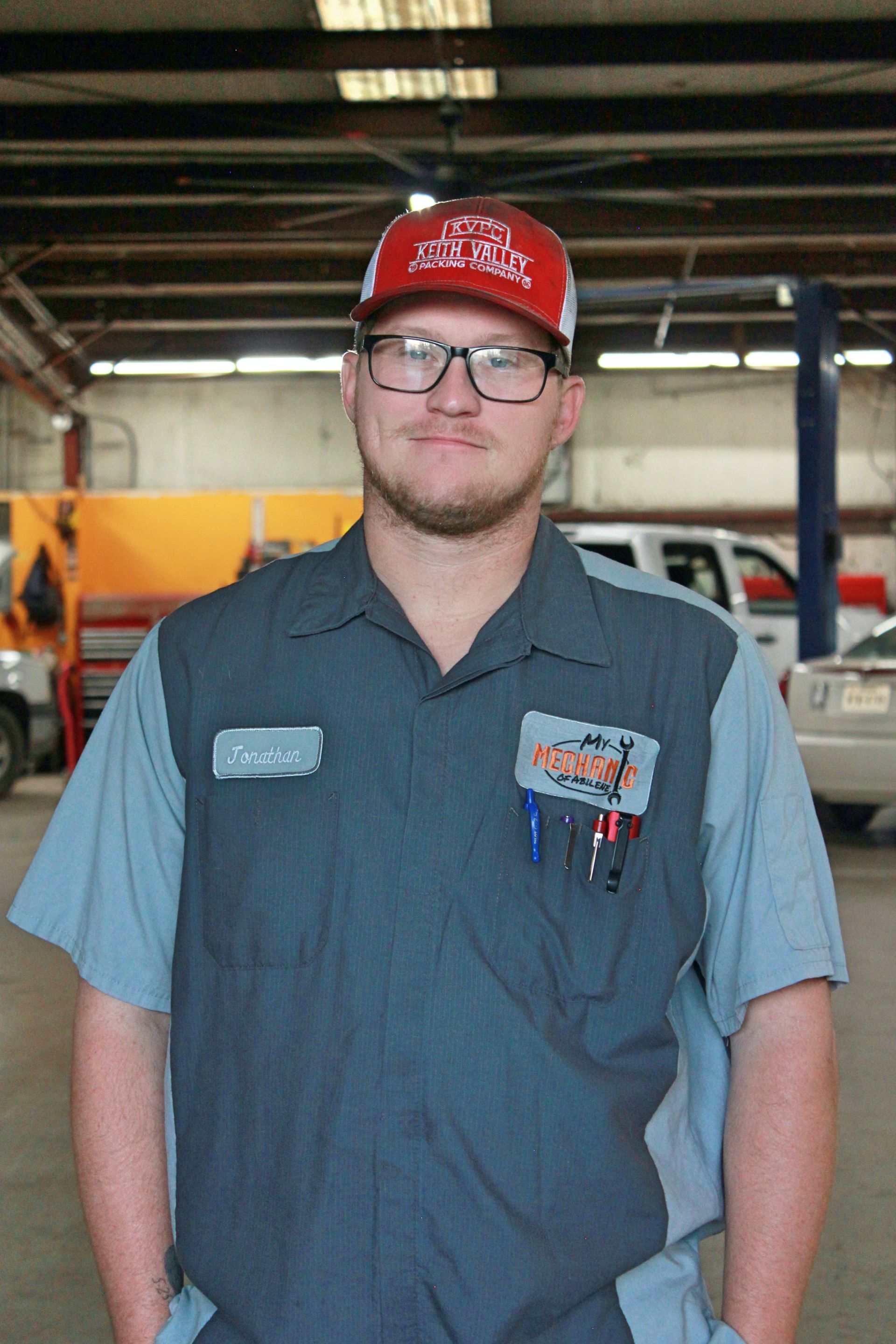 Mechanic in a shop, wearing glasses and a red hat, smiles at the camera, hands in pockets.