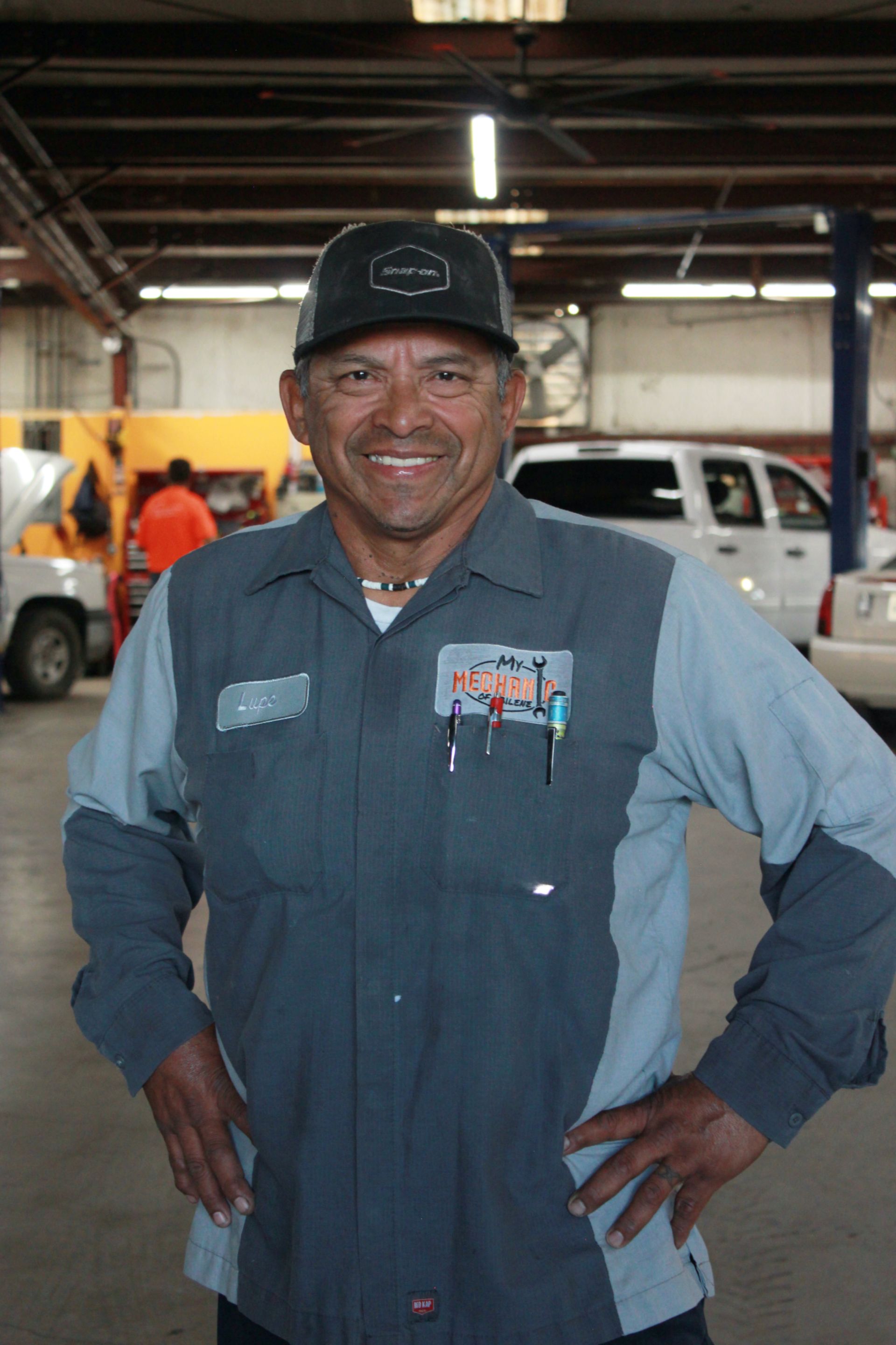 Man in mechanic's uniform smiles, hands on hips, in a garage setting with cars.