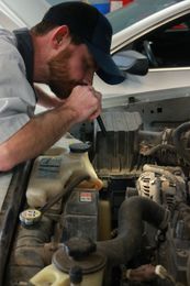 Mechanic inspecting engine compartment with a flashlight. He's wearing a blue cap, working inside a garage.