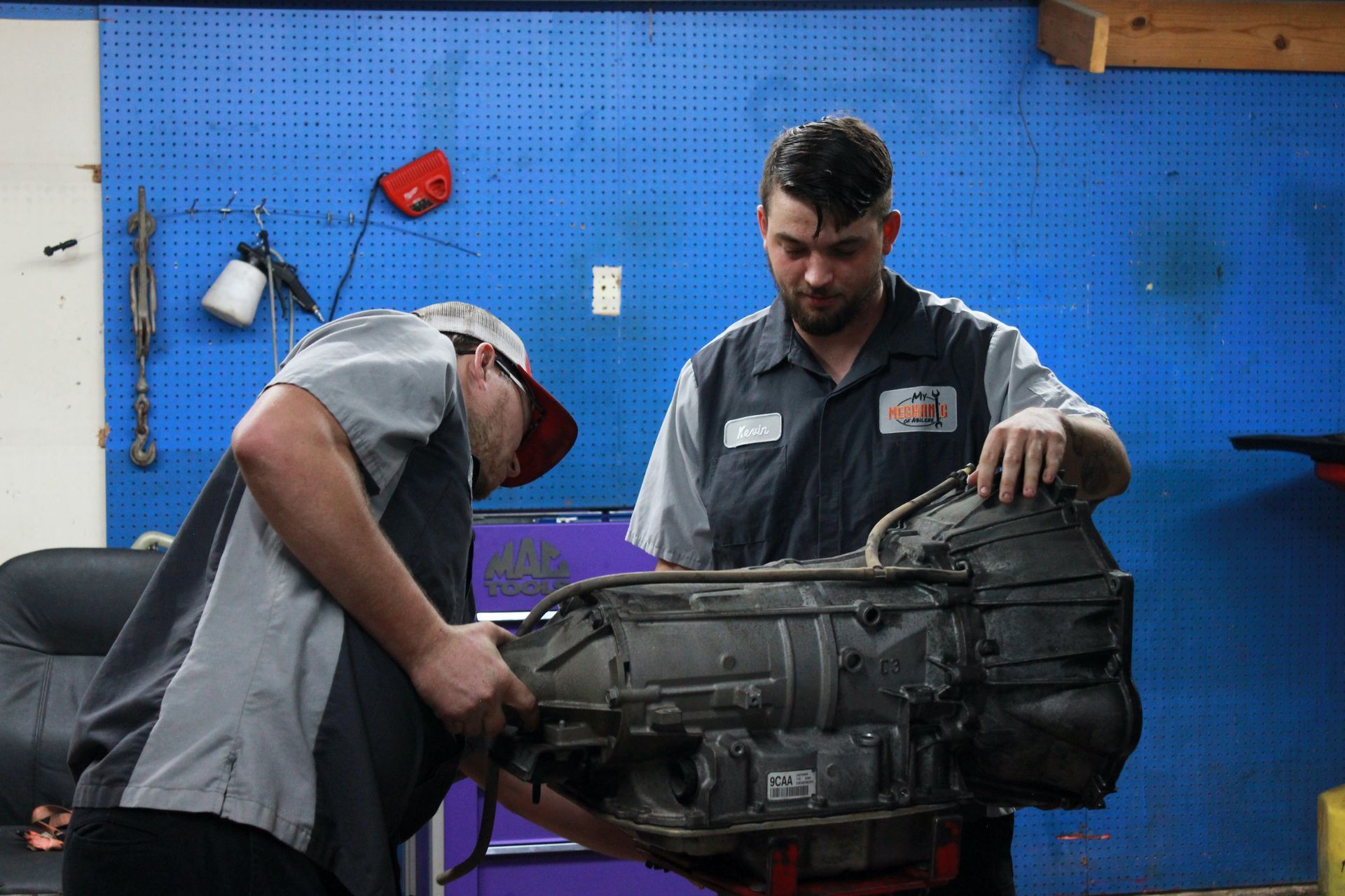 Two mechanics working on a transmission in a garage with blue walls.