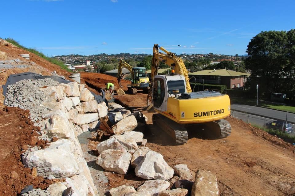 A Yellow Excavator Is Driving Down A Dirt Road Next To A Pile Of Rocks — Australian Rock Walls In Burleigh Heads, QLD