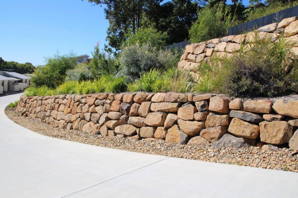A Stone Wall Is Surrounded By Trees And Bushes On The Side Of A Road — Australian Rock Walls In Burleigh Heads, QLD