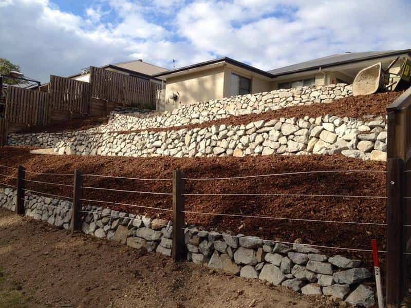 Stone Retaining Walls With Mulch, Wooden Fence, And A House On A Hillside