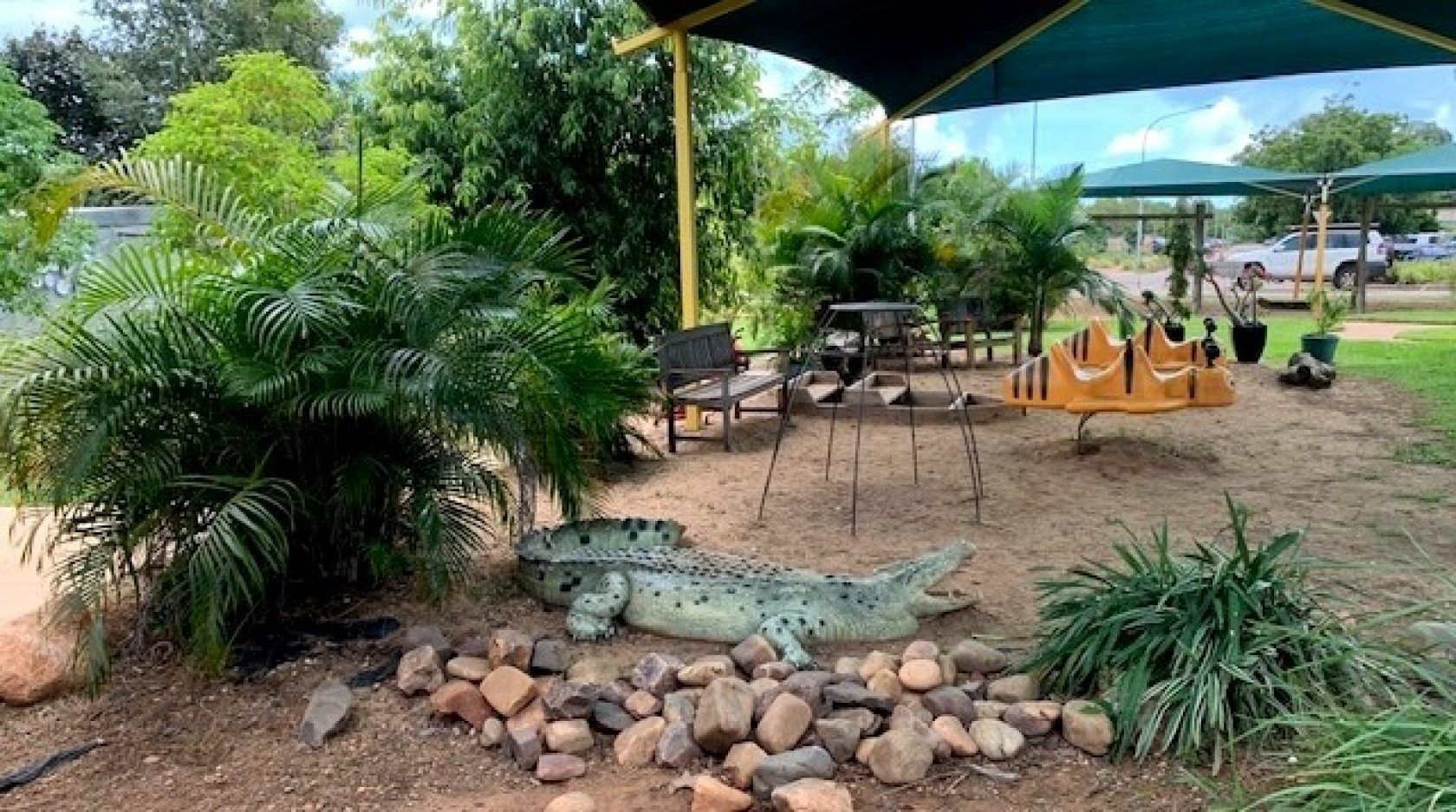 A crocodile statue rests near rocks in a park with shade sails, greenery, and a table.