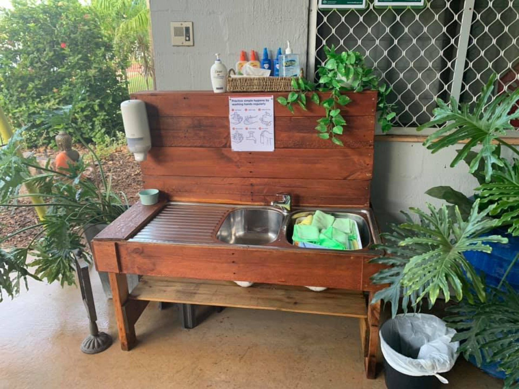 Wooden outdoor handwashing station with soap, water, and instructions near plants and building.