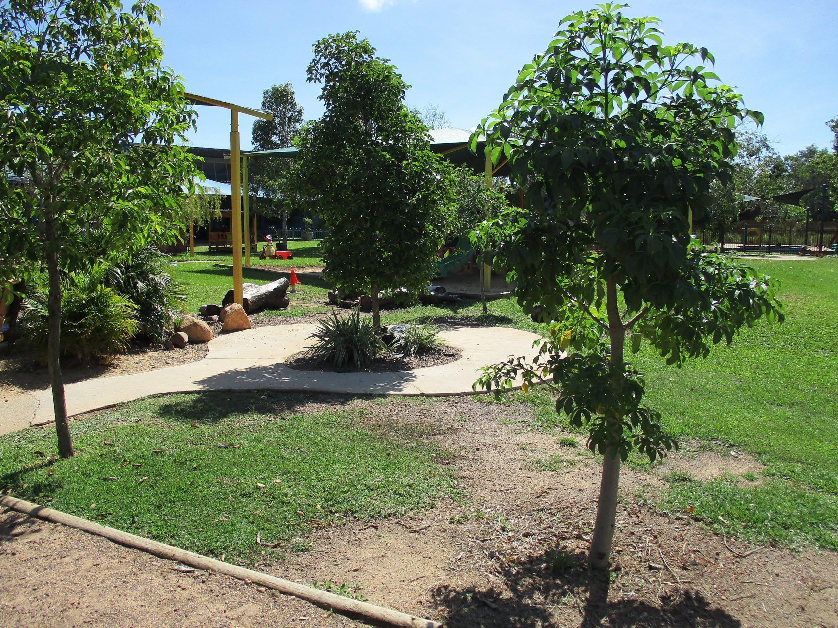 Park with trees, path, grass, and a playground in the background under a sunny sky.