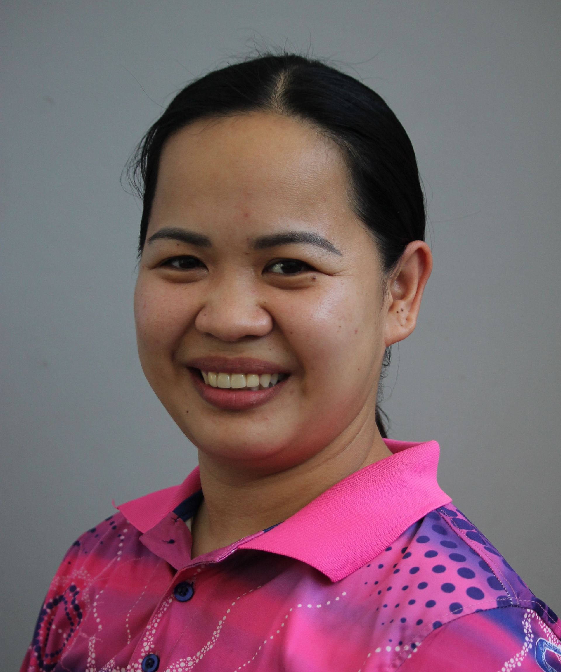 Smiling woman with dark hair wearing a pink patterned shirt, against a grey backdrop.