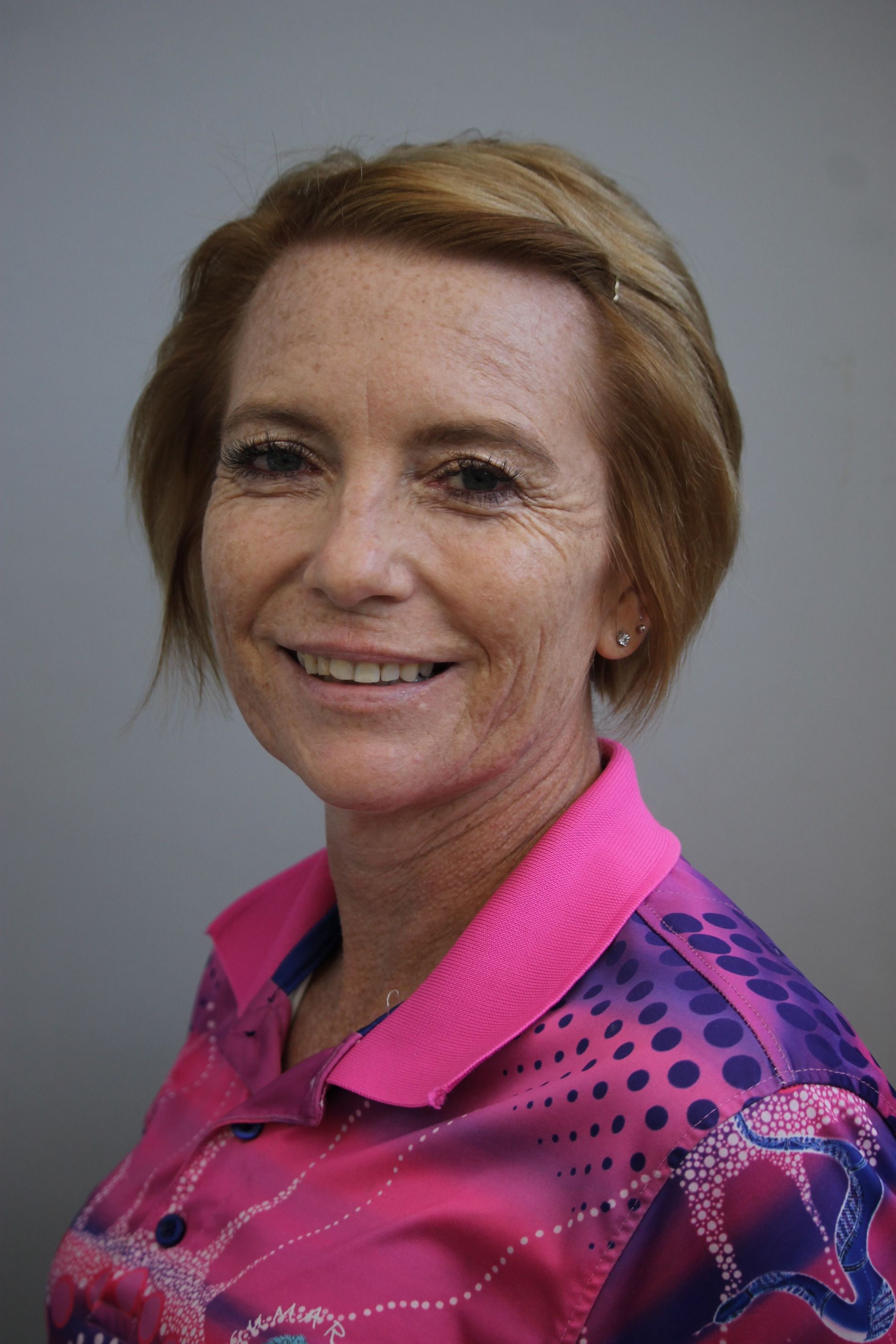 Woman with short auburn hair, smiling, wearing a pink and purple patterned shirt against a gray background.