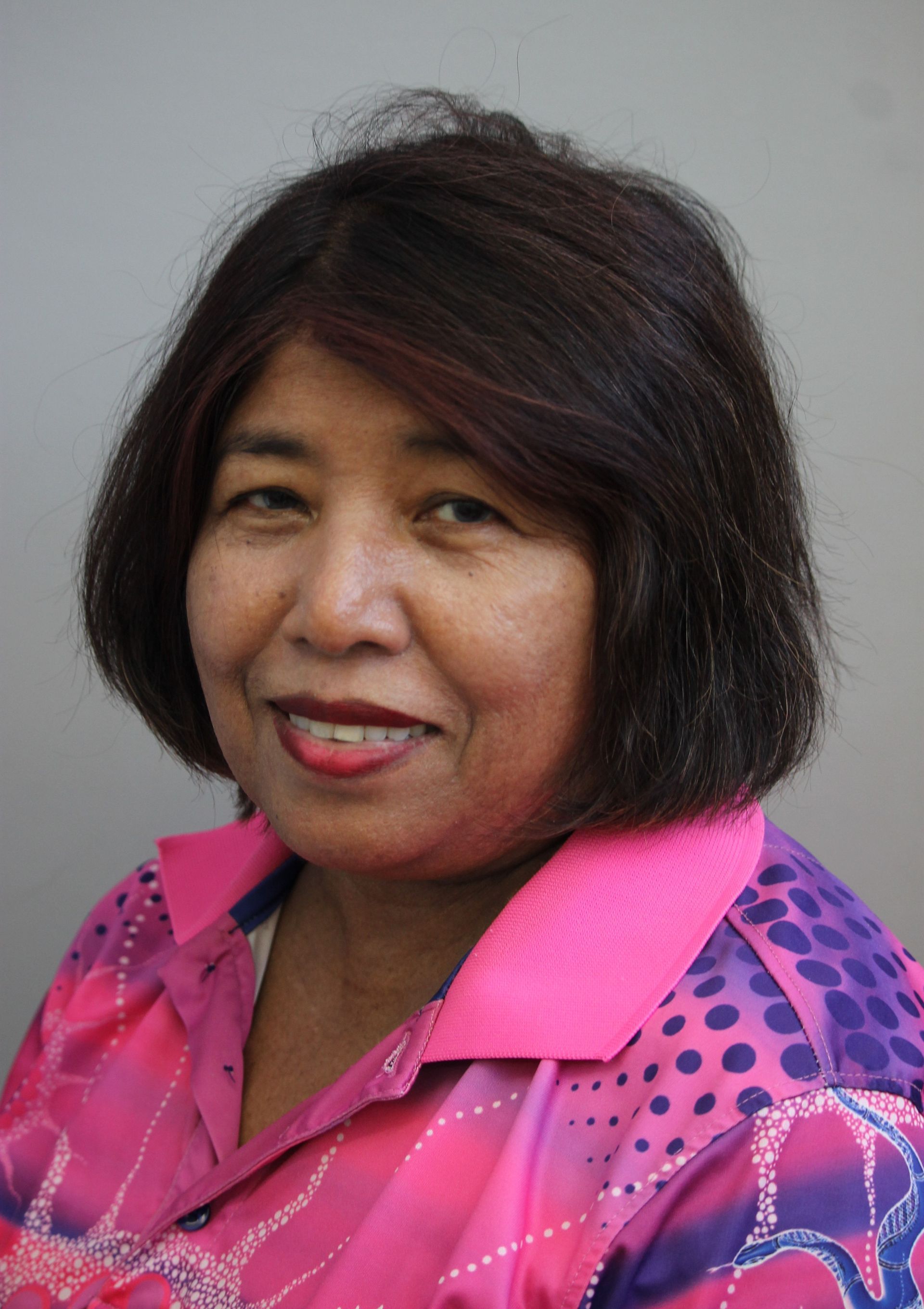 Woman with dark hair, wearing a pink patterned shirt, smiling.