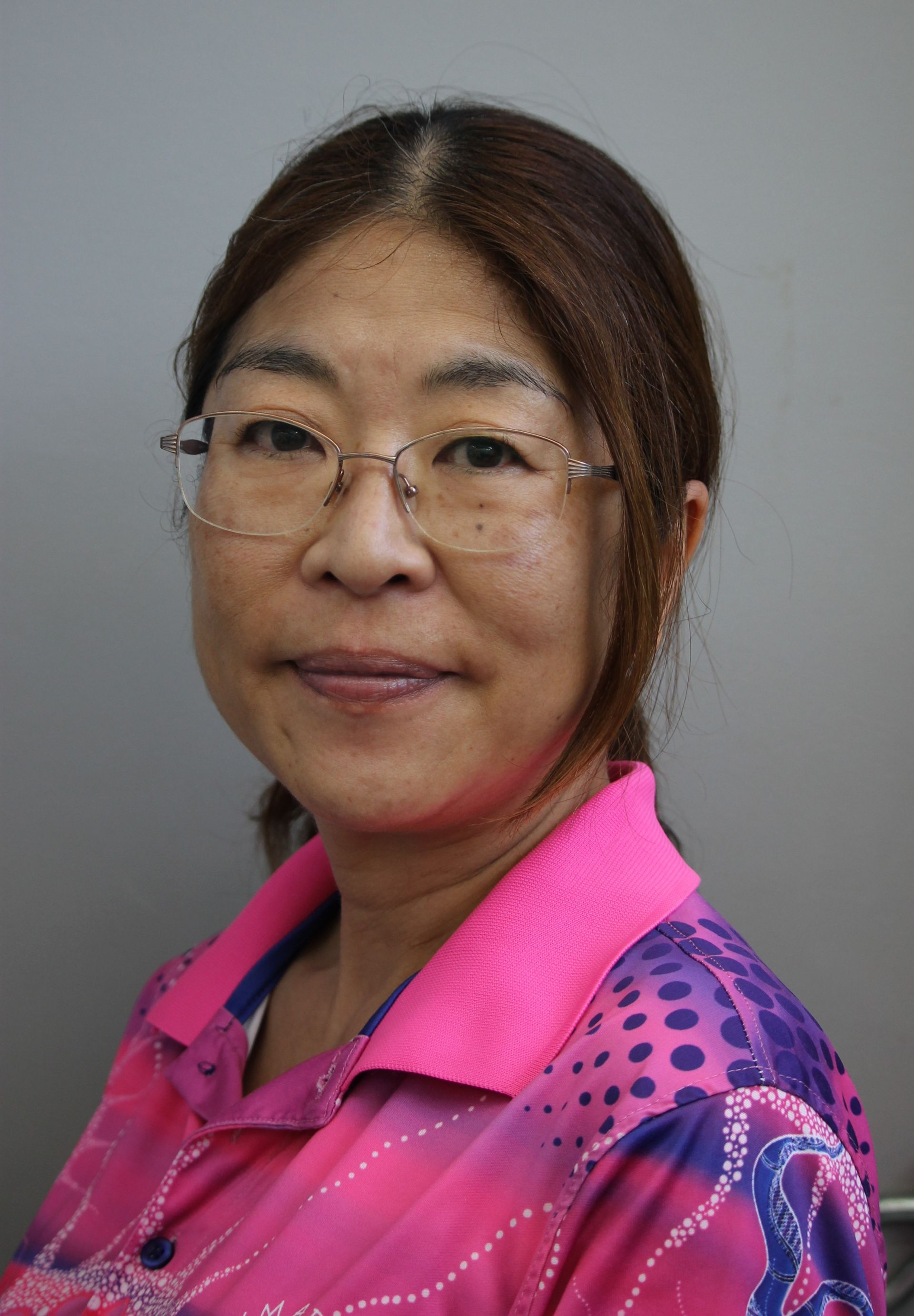 Woman with glasses, wearing a pink patterned shirt, smiles at the camera against a gray background.