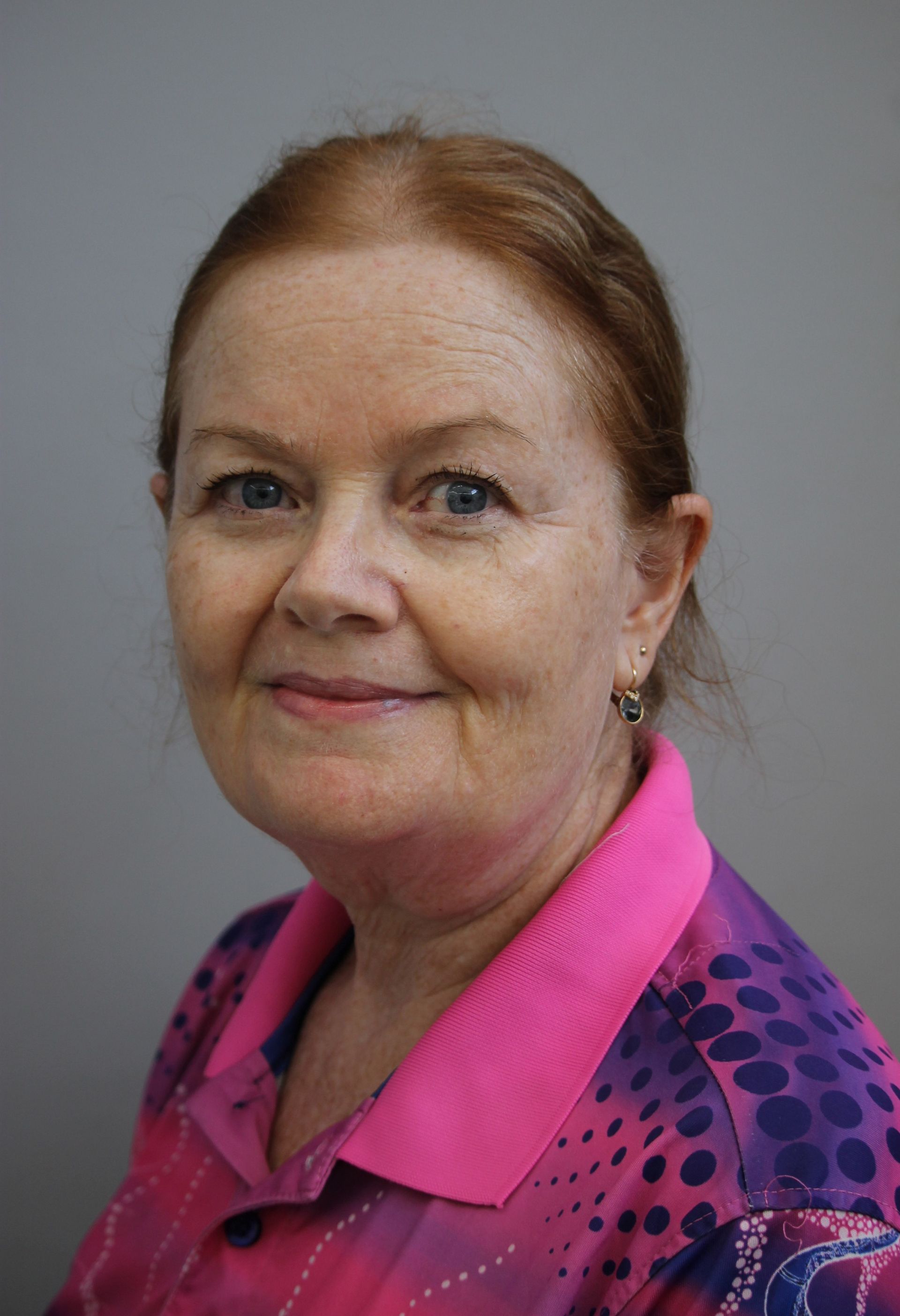 Woman with red hair, smiling, wearing a pink and purple shirt, in front of a gray background.