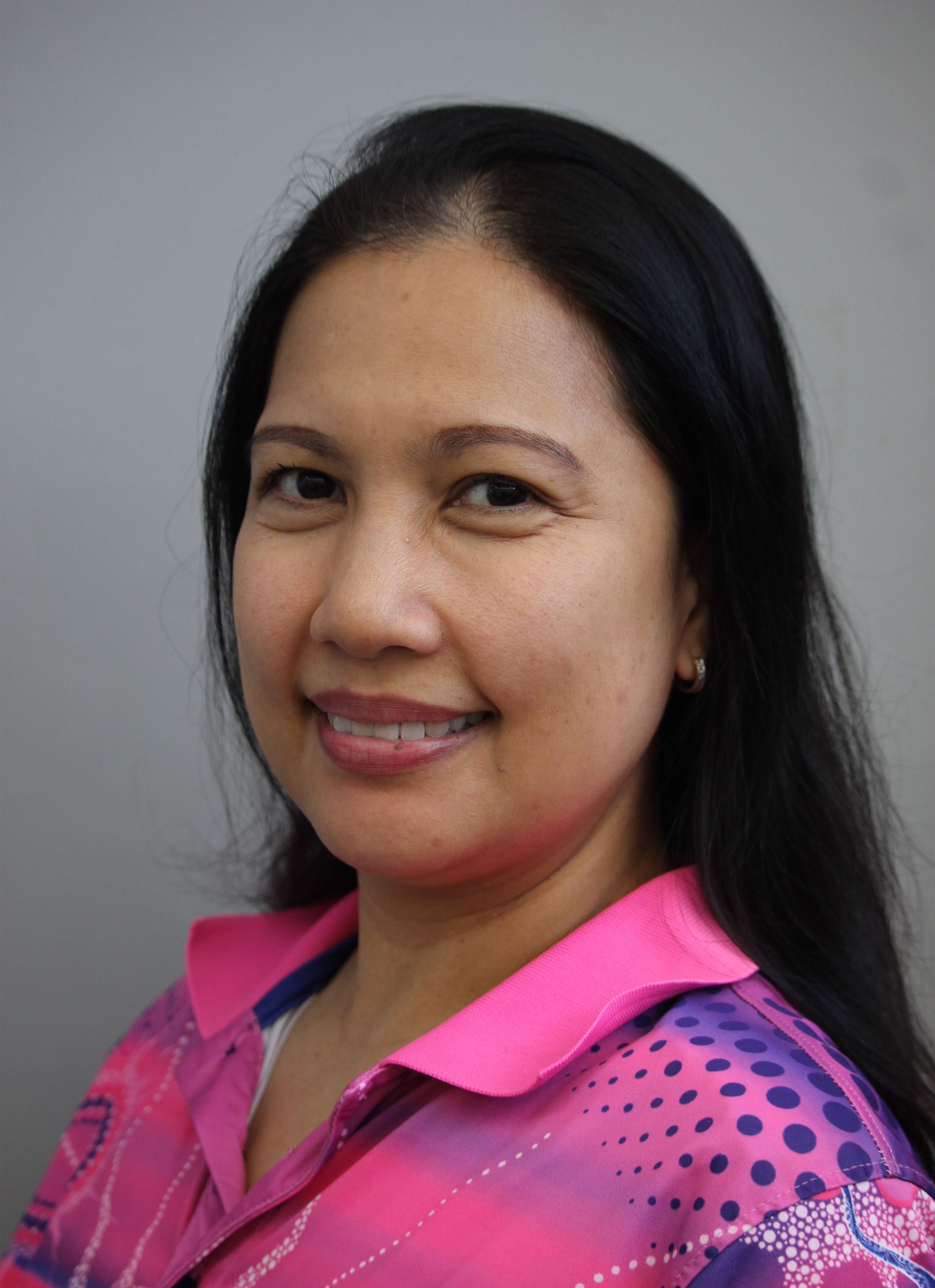 Woman with dark hair smiling, wearing a pink patterned shirt against a grey background.