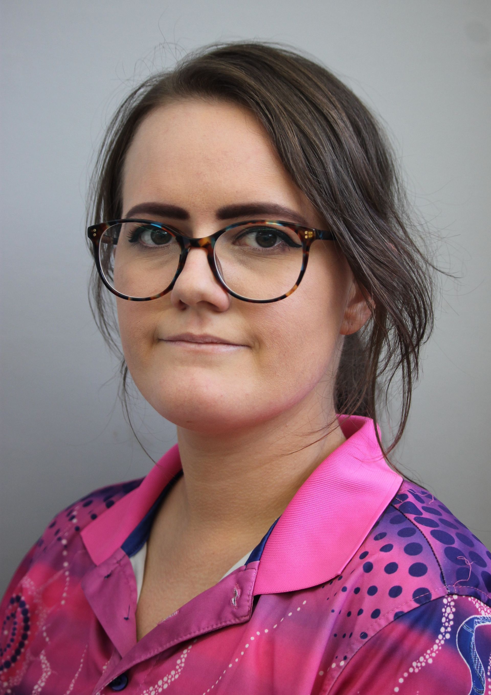 Woman with glasses wearing a pink patterned shirt, facing the camera with a neutral expression.