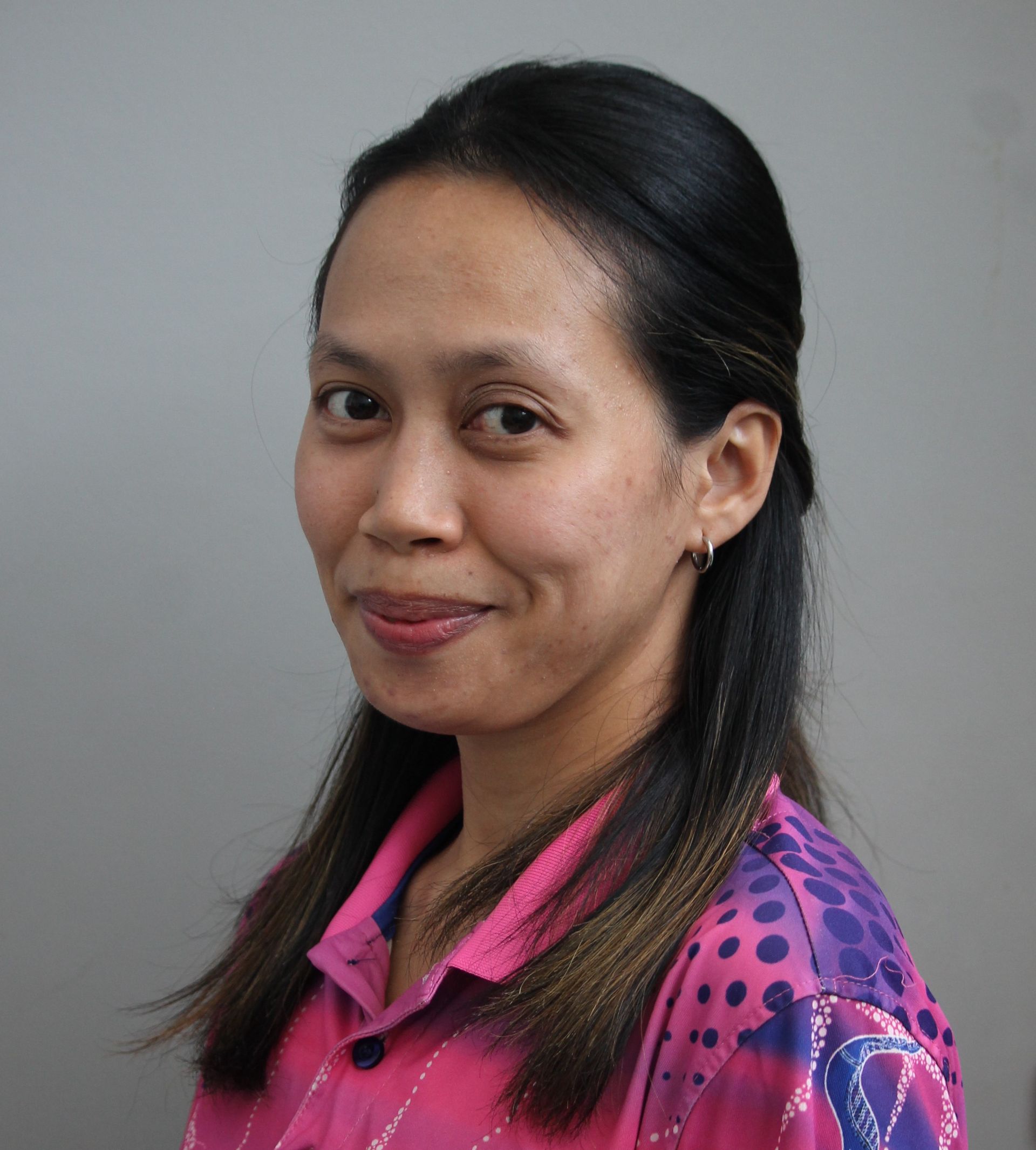 Woman with dark hair, wearing a pink patterned shirt, smiles at the camera against a gray backdrop.