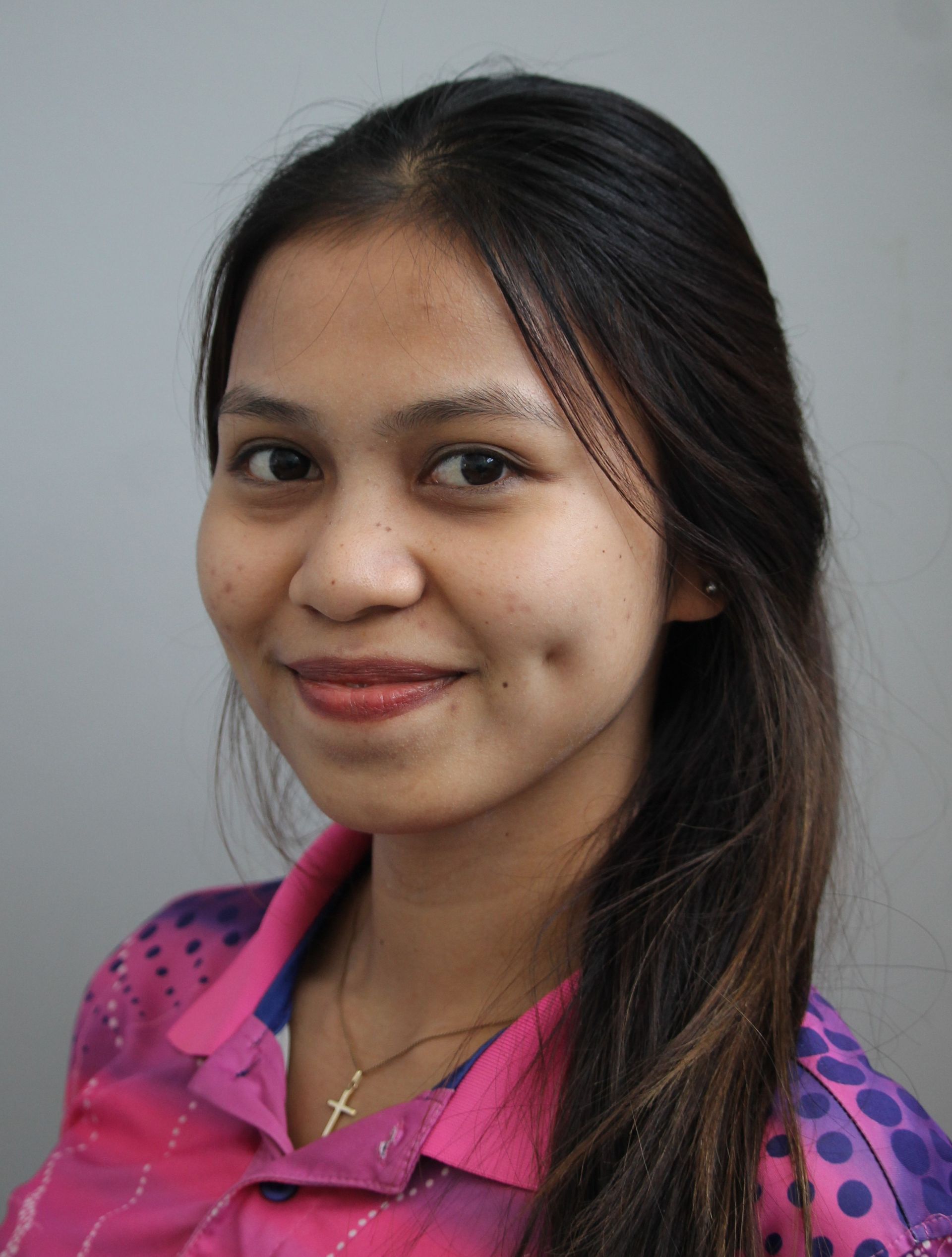 Woman with long dark hair, smiling, wearing a pink shirt with a pattern, set against a gray background.