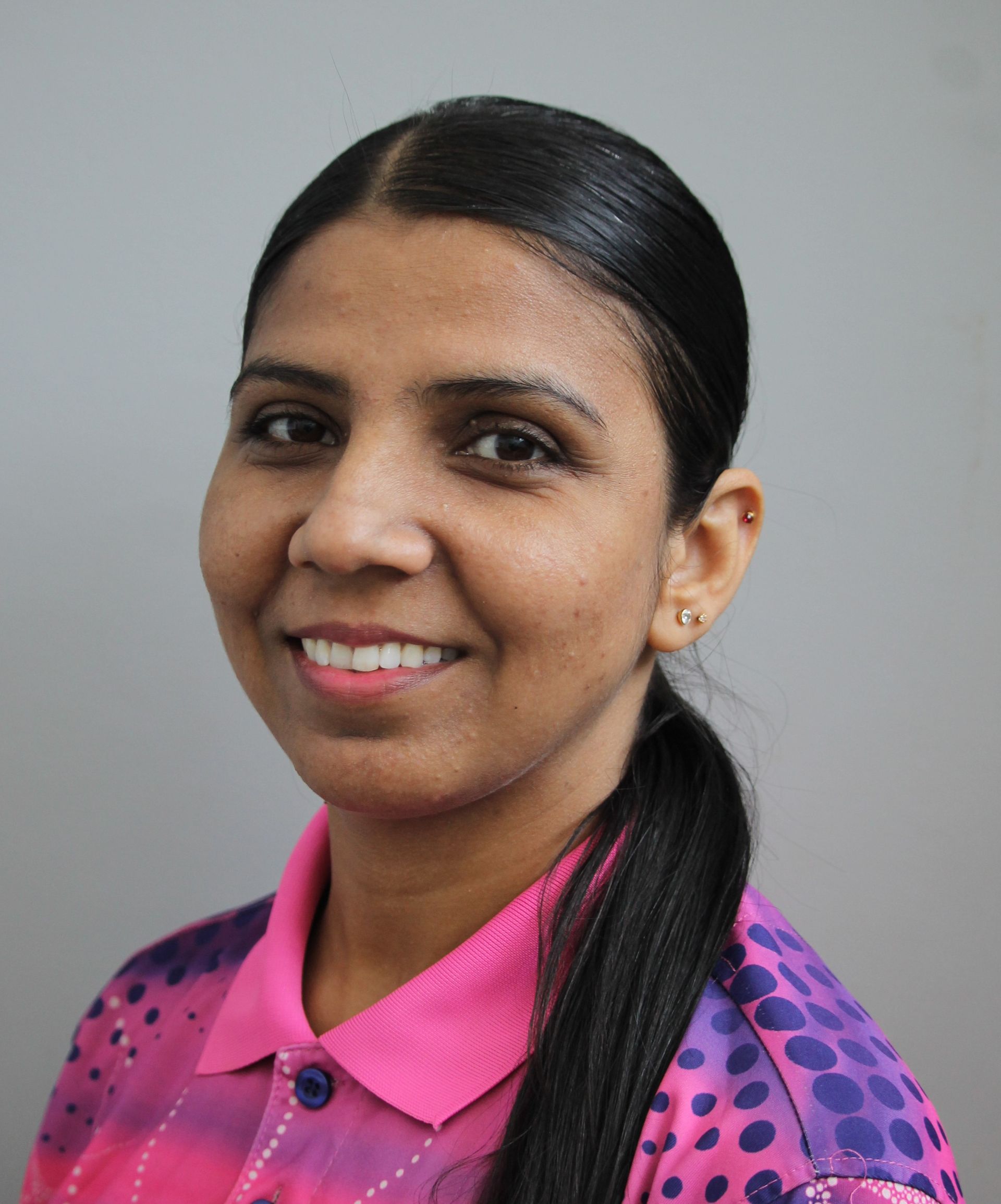 Woman with dark hair in a ponytail smiling, wearing a pink and purple patterned collared shirt.