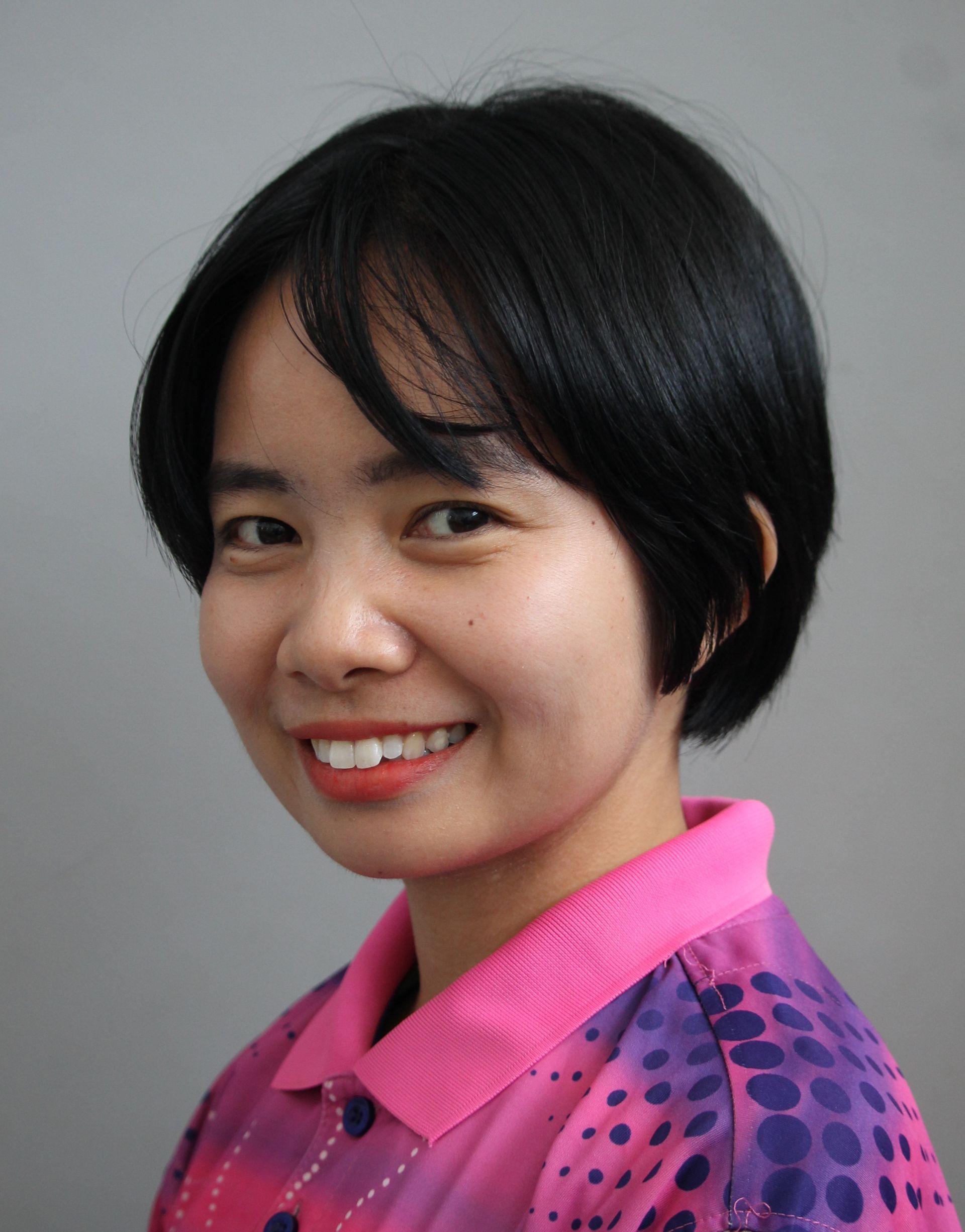 Woman with short black hair smiles, wearing a pink and purple shirt, against a gray background.