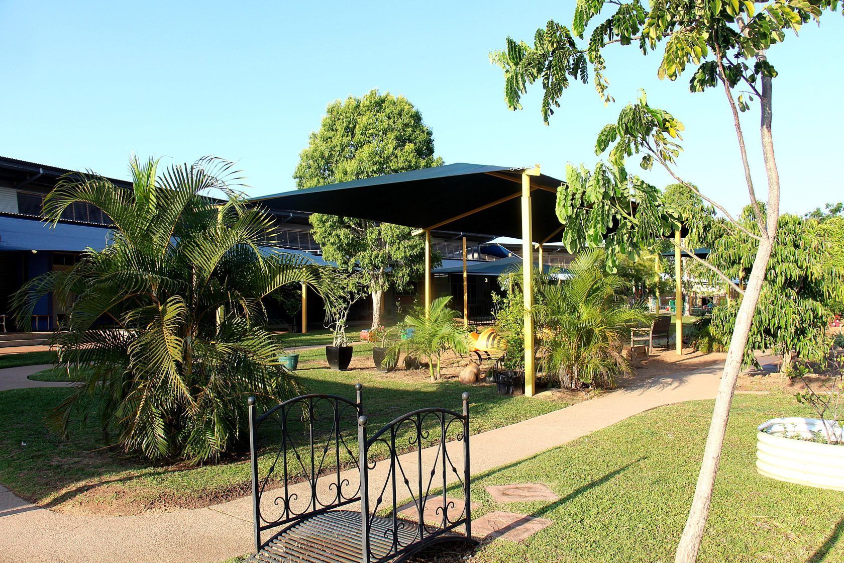 Lush garden scene with a covered seating area, pathways, and greenery under a blue sky.