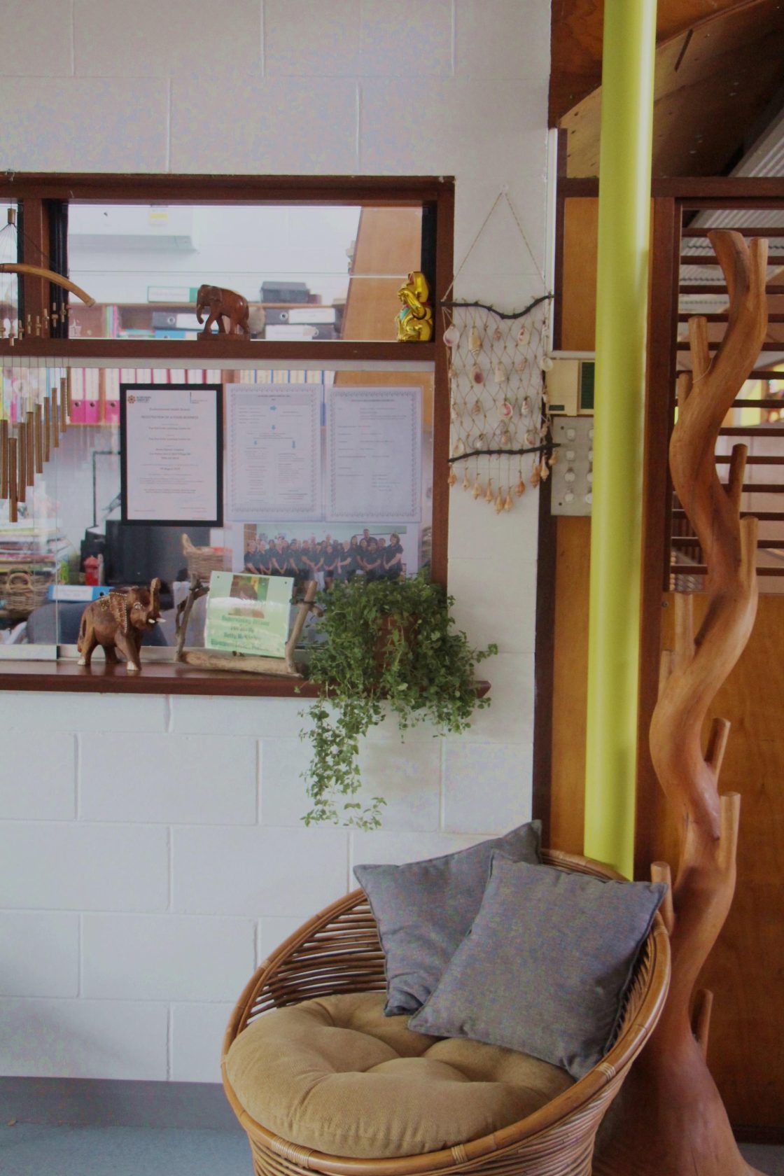Wicker chair with cushions by a wall with a window, plants, and decorative wood sculpture.