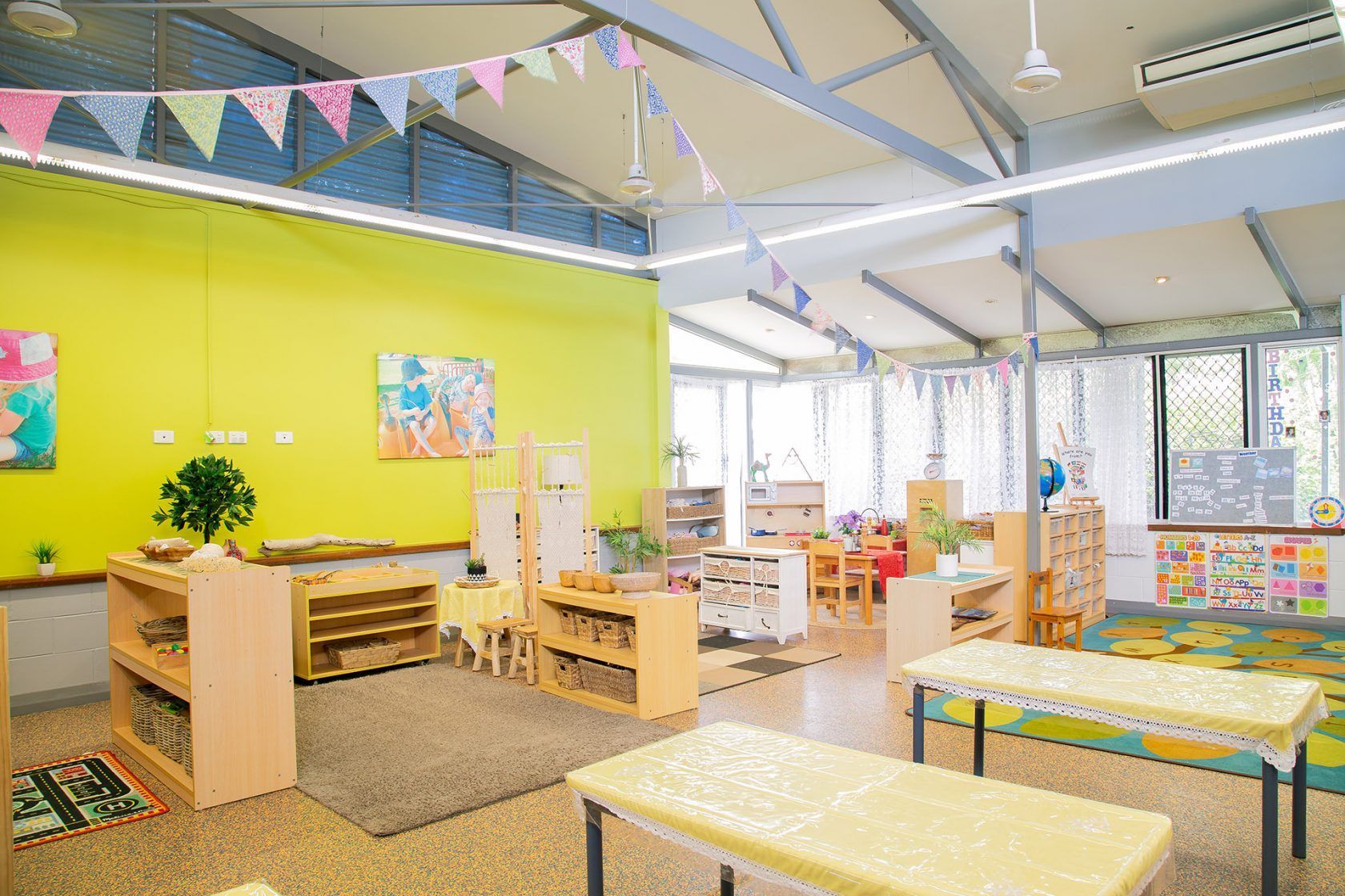 Kindergarten classroom with wooden furniture, green wall, and colorful decorations.