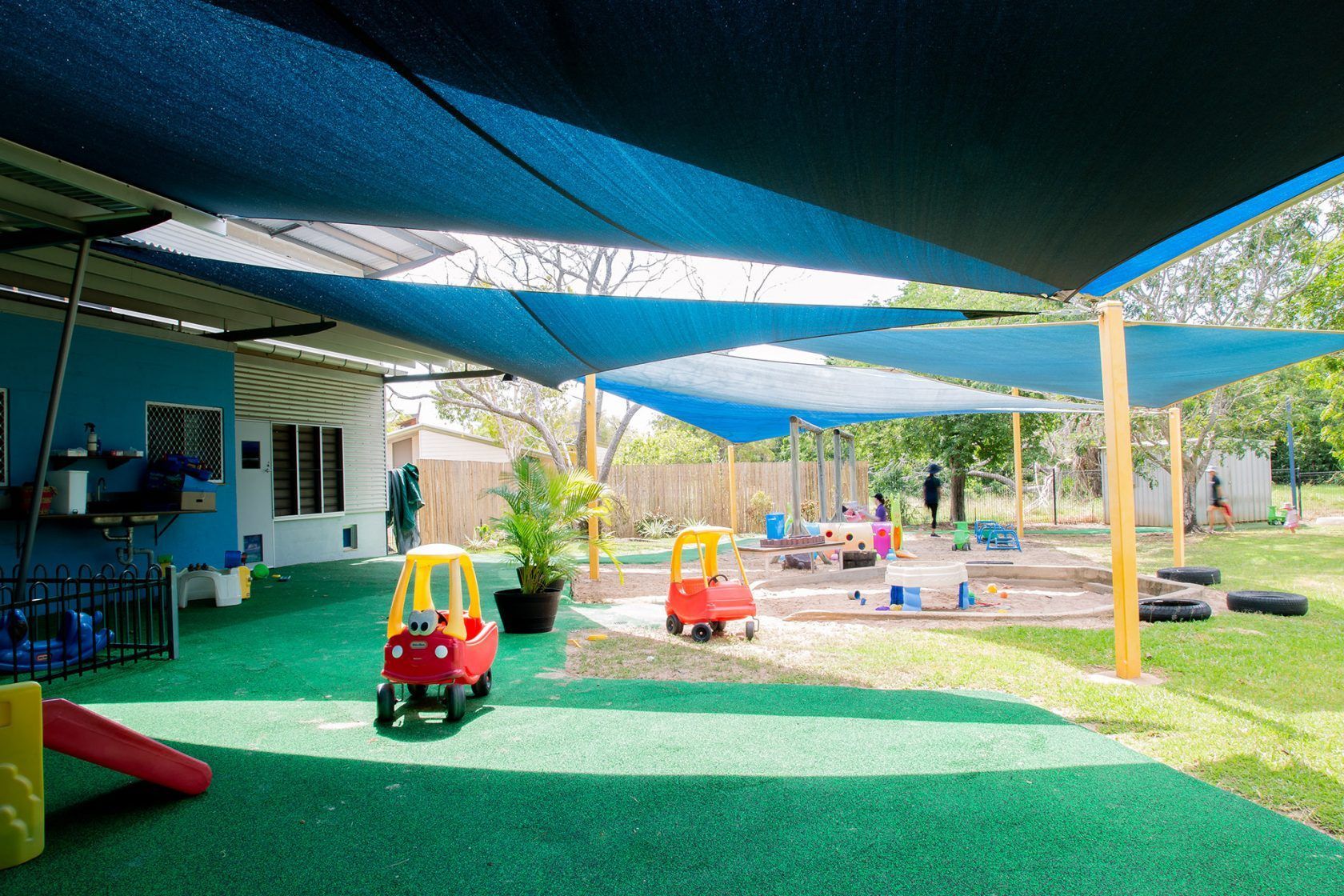 Children's playground with shade sails, green turf, toy cars, and sandboxes under a blue sky.